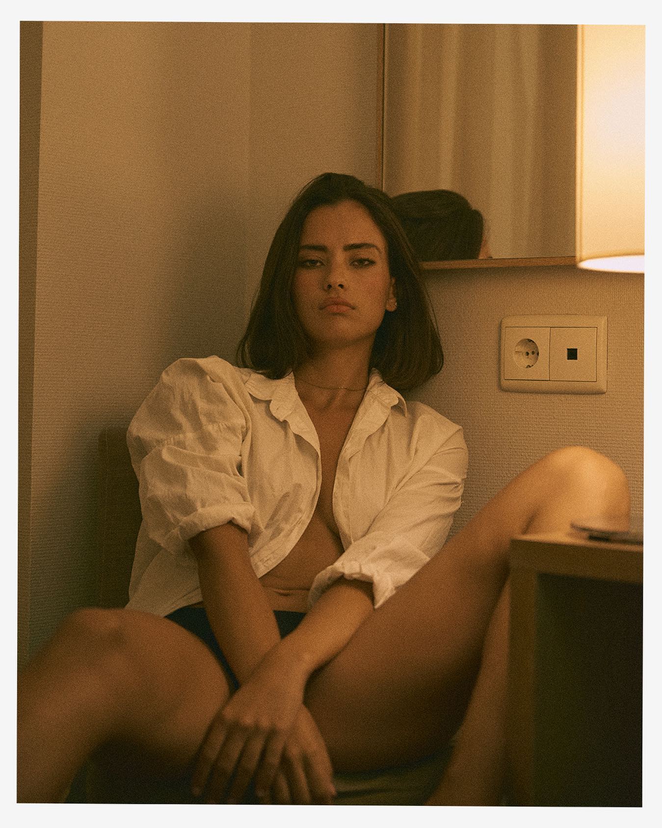 Modelo femenina sentada en una habitación con luz cálida, vistiendo una camisa blanca desabotonada. Fotografía editorial con un estilo íntimo y minimalista, capturada en un entorno interior con iluminación suave.